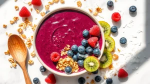 Vibrant overhead shot of a deconstructed acai bowl showing the deep purple frozen acai base, scattered granola clusters, fresh raspberries, blueberries, sliced kiwi, coconut flakes, and a wooden spoon on a light marble surface with natural sunlight