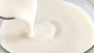 Close-up photograph of fresh heavy cream being poured into a clear glass bowl, showing rich white liquid texture and glossy surface, professional kitchen lighting, shallow depth of field highlighting cream consistency